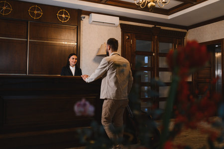 Man arriving at classic hotel lobby, checking in with smiling female receptionist. Hospitality service and travel accommodationの写真素材