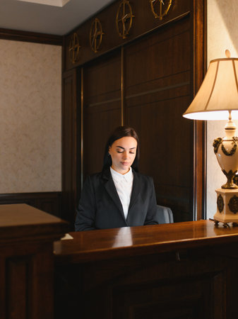Professional woman standing at a dark wood reception desk, providing hospitality service in a hotel lobby interiorの写真素材
