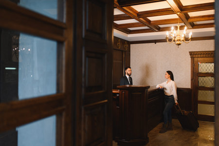 Hotel receptionist assisting a woman guest with luggage at a dark wooden reception desk in a classic lobby interiorの写真素材