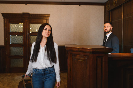 Woman arriving at a classic hotel lobby, pulling a suitcase while a male receptionist stands behind the front deskの写真素材