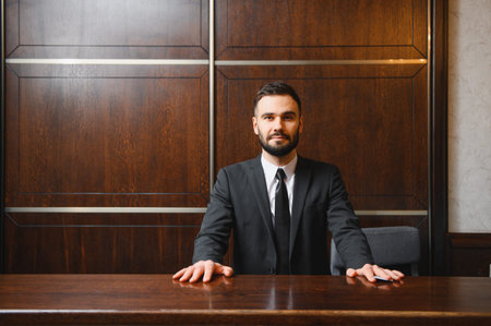 Professional male receptionist standing behind a wooden front desk in a luxury hotel lobby, providing hospitality serviceの写真素材