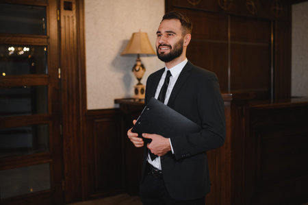 Smiling man in a suit holding a clipboard, standing at a reception desk in a hotel lobby, portraying hospitalityの写真素材