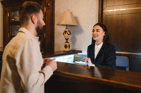 Young man checking in at a hotel reception, talking with a friendly female concierge providing excellent hospitality serviceの写真素材