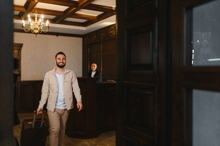 Smiling man pulling luggage, entering a hotel lobby as a friendly receptionist waits behind the counterの写真素材
