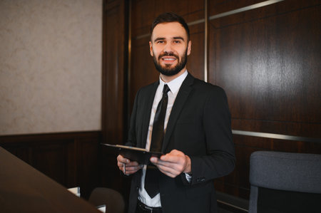 Friendly male receptionist standing at a hotel lobby desk, holding a clipboard and pen, ready to assist guestsの写真素材