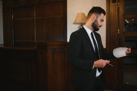 Professional man wearing a suit and tie, reviewing paperwork in a hotel reception area. Concept of business travel and administrationの写真素材