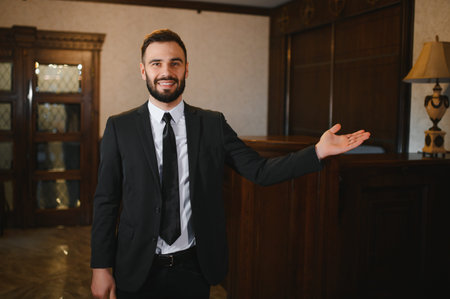 Friendly male receptionist wearing a suit, smiling and gesturing for welcome or showing direction in a hotel lobbyの写真素材