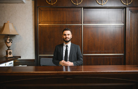 Smiling man in a suit working at a hotel reception desk, providing hospitality service to guests in a elegant lobbyの写真素材