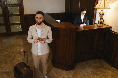 Smiling guest standing in a classic hotel lobby with a suitcase, holding a phone, waiting for receptionist serviceの写真素材
