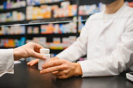 Pharmacist standing behind a protective screen, wearing a white coat, giving a prescription bottle to a customerの写真素材