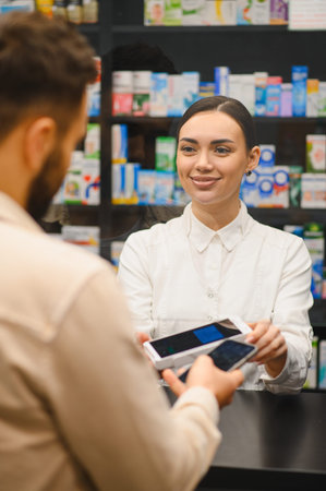 Pharmacist holding a payment terminal for a customer paying with a smartphone in a modern pharmacy with medicine shelvesの写真素材