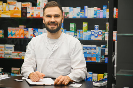 Male pharmacist in white coat smiling at camera, sitting at the counter with prescription pad in a pharmacyの写真素材