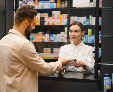 Young man using a mobile phone for contactless payment at a pharmacy counter, interacting with a smiling female pharmacistの写真素材