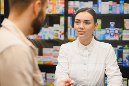 Pharmacist working behind counter, smiling while handing medicine to a male customer, providing consultation and healthcare serviceの写真素材