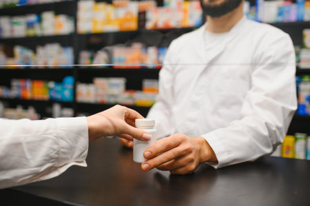 Pharmacist handing a medicine bottle to a customer across a protective screen at a modern pharmacy counter, symbolizing healthcare and serviceの写真素材