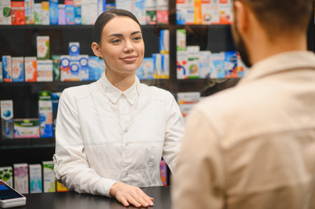 Friendly female pharmacist wearing white coat, smiling and helping a customer with medication or advice inside a modern drugstoreの写真素材