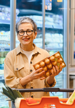 Smiling mature woman shopping for groceries, holding a carton of eggs in a supermarket aisle with her shopping cartの写真素材
