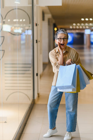 Mature woman with gray hair wearing glasses, holding shopping bags, showing great excitement near a store windowの写真素材
