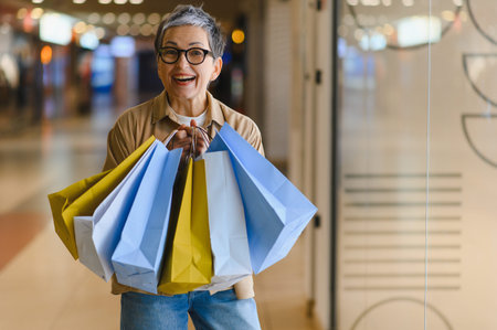 Cheerful mature woman smiling, holding multiple colorful shopping bags, walking in a modern mall aisleの写真素材