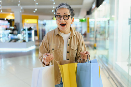 Senior woman walking in a mall, holding several shopping bags, expressing excitement and happiness with consumerismの写真素材