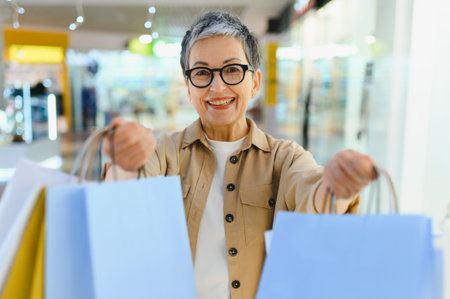 Happy senior woman shopping in a mall, holding several colorful bags, smiling at camera. Consumerism...