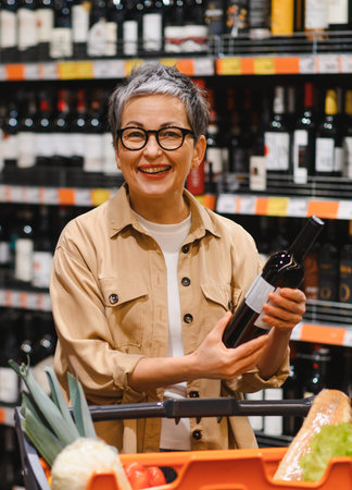 Happy mature woman holding a wine bottle, smiling brightly while grocery shopping in a supermarket aisleの写真素材