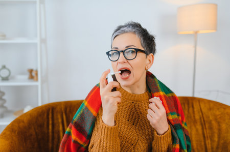 Woman feeling unwell at home, sitting on sofa, using throat spray for sore throat or other illness...