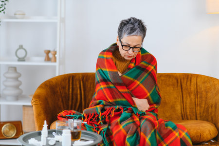 Senior woman suffering from cold or flu, wrapped in a blanket on a couch, experiencing fever and shiveringの写真素材