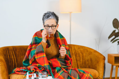Woman wrapped in a blanket on a couch, experiencing flu or cold symptoms, finding comfort with a warm drinkの写真素材