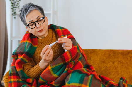 Senior woman feeling unwell with a cold or flu, checking her body temperature with a thermometer...