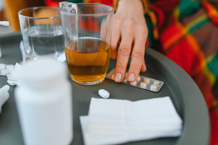 Hand reaching for pills on a tray with tea and glass of water, managing flu or cold symptoms at home during recovery routineの写真素材