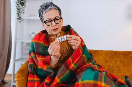 Elderly woman experiencing sickness, wrapped in a blanket on the sofa and holding a blister pack of medicationの写真素材