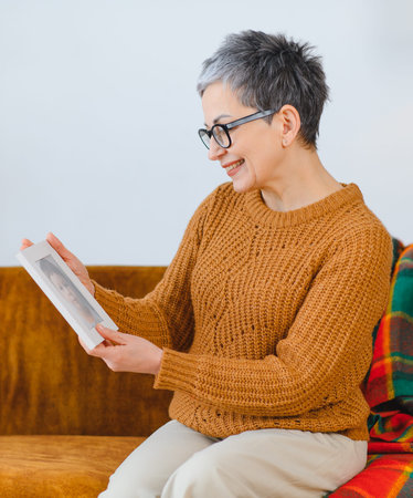 Senior woman at home smiling, holding a framed picture, remembering special moments and cherishing past memoriesの写真素材