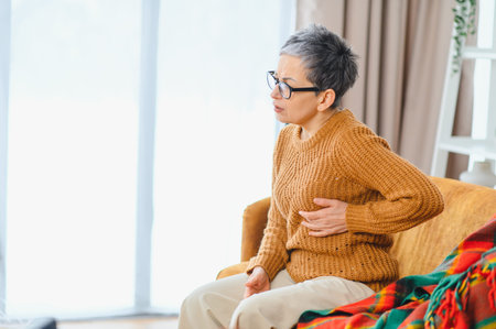 Woman sitting on sofa, clutching her chest in pain. Experiencing symptoms of heart attack, shortness...