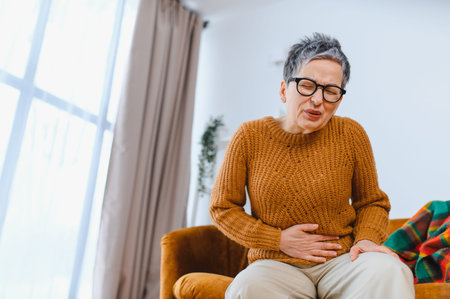 Senior woman suffering from abdominal ache, experiencing discomfort and indigestion while sitting on...