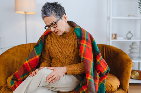 Elderly woman sitting on a sofa, rubbing her aching knee, feeling discomfort and joint pain due to arthritisの写真素材