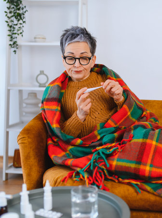 Older woman checking thermometer, feeling unwell at home. Wrapped in blanket fighting, illness with...