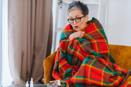 Senior woman feeling unwell with flu or cold symptoms, wrapped in a warm plaid blanket, staying home to recoverの写真素材