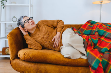 Senior woman suffering from severe abdominal pain, lying on a couch with a hand on her stomach. Health problemの写真素材