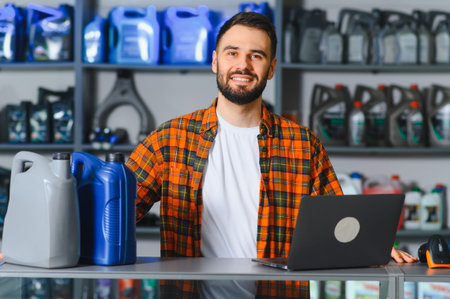 Young male auto parts shop owner smiling behind counter with motor oil on shelves and laptop,...