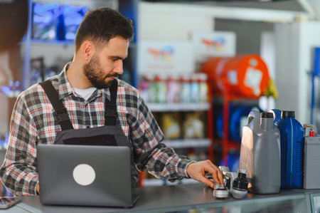 Male mechanic in uniform using laptop, choosing car parts, fluids, and components at an automotive...