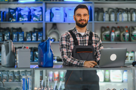 Young auto mechanic working in workshop, using laptop for orders and inventory, standing near...