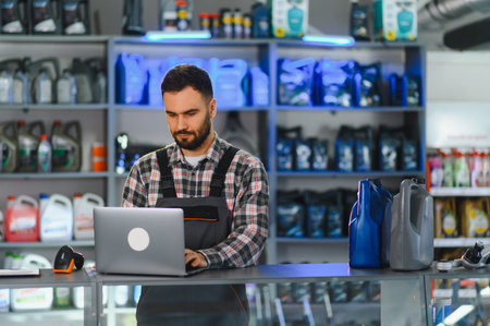 Auto service technician using laptop at counter in car maintenance shop, surrounded by motor oil and...