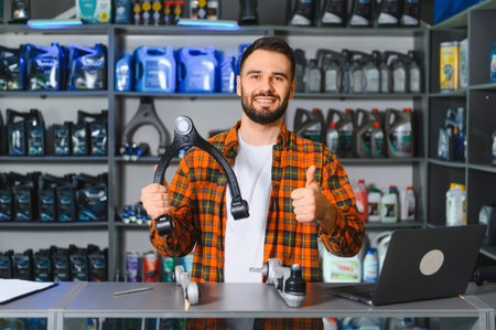 Bearded auto parts specialist at counter holding a control arm and giving thumbs up, smiling confidently in parts store environmentの写真素材