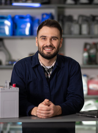 Male automotive technician smiling while standing at desk with car battery and laptop in...