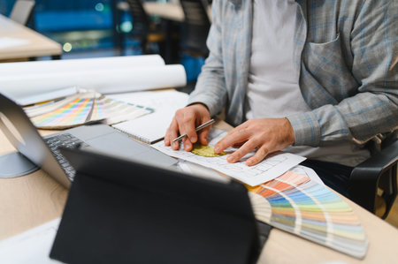 Interior designer hands working with an architectural blueprint, color palette samples, and a laptop...