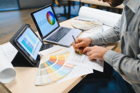 Designer's hands drawing on architectural plans with laptop, tablet, and color palette, creating new home or office spaceの写真素材
