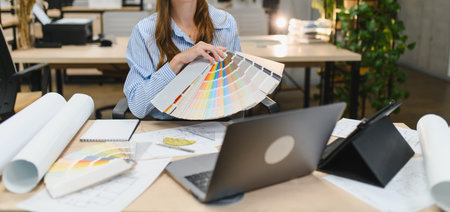 Designer choosing colors from a swatch book, working on a laptop with blueprints for an interior...