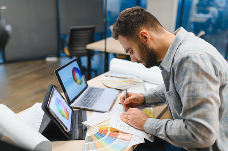 Creative professional man working in office, using laptop, tablet, and color swatches for...