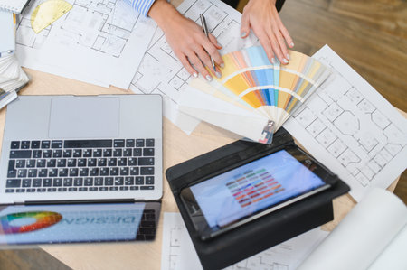 Hands of a designer holding color swatches for selecting interior design palette, with floor plans, laptop, and tablet on a deskの写真素材